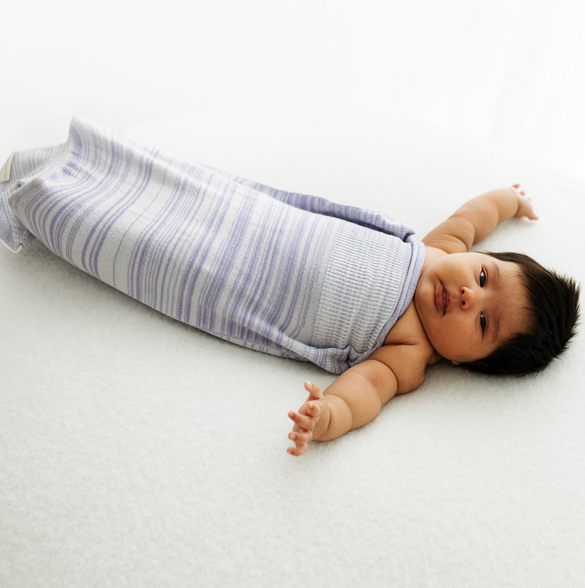 A baby lying in a lavender swaddle with its arms out.