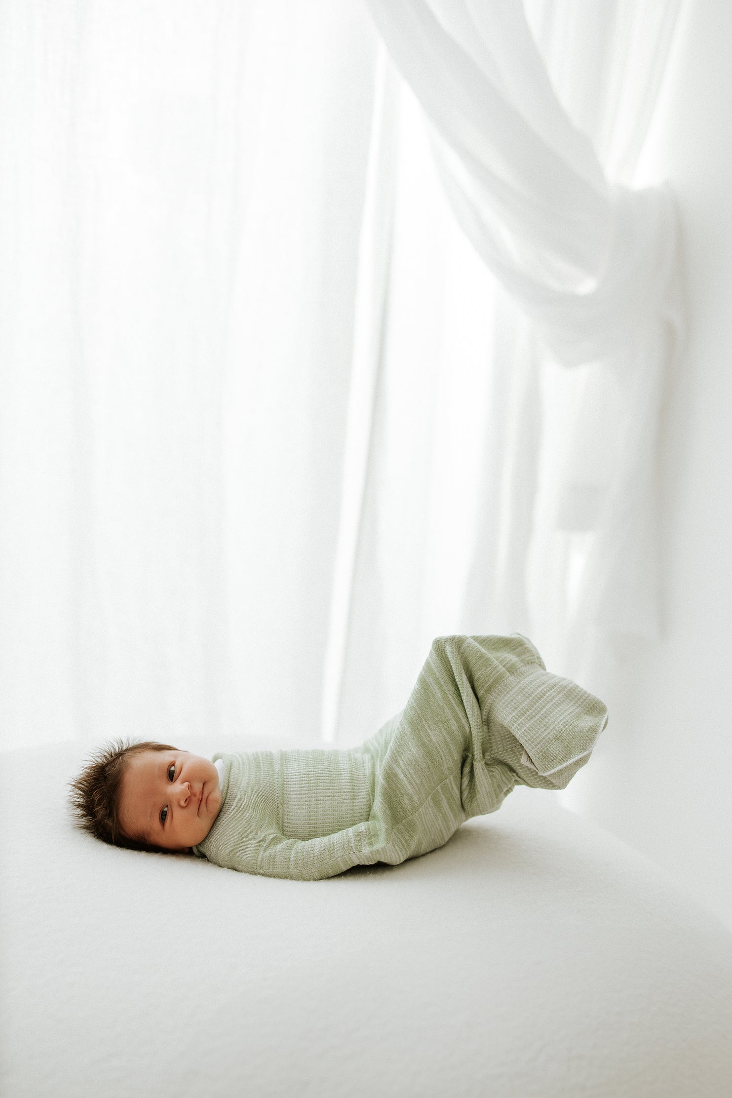 A baby lying in a mint colored sleep sack swaddle.